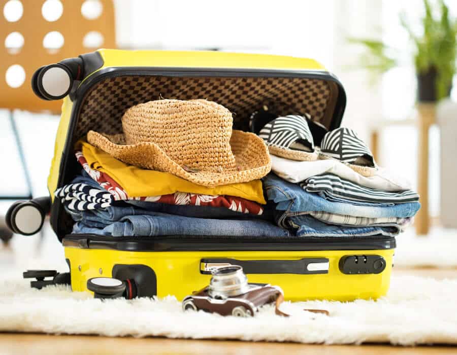 Open yellow suitcase filled with clothes, a straw hat, and sandals on a soft surface.