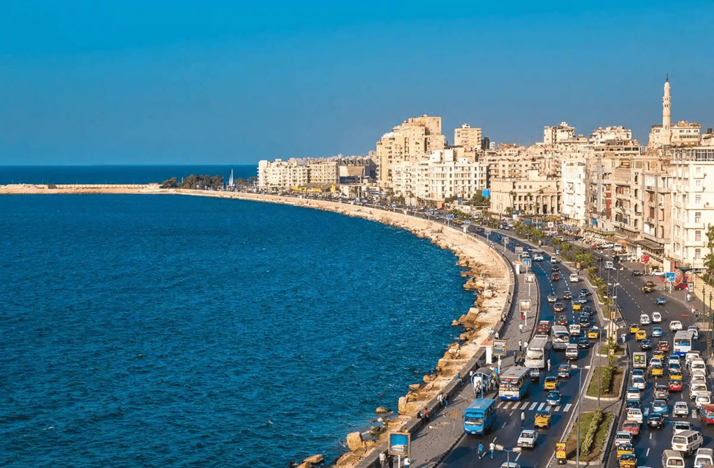 A high-angle view of the curved Corniche road and waterfront in Alexandria, Egypt, with blue sea and cityscape.